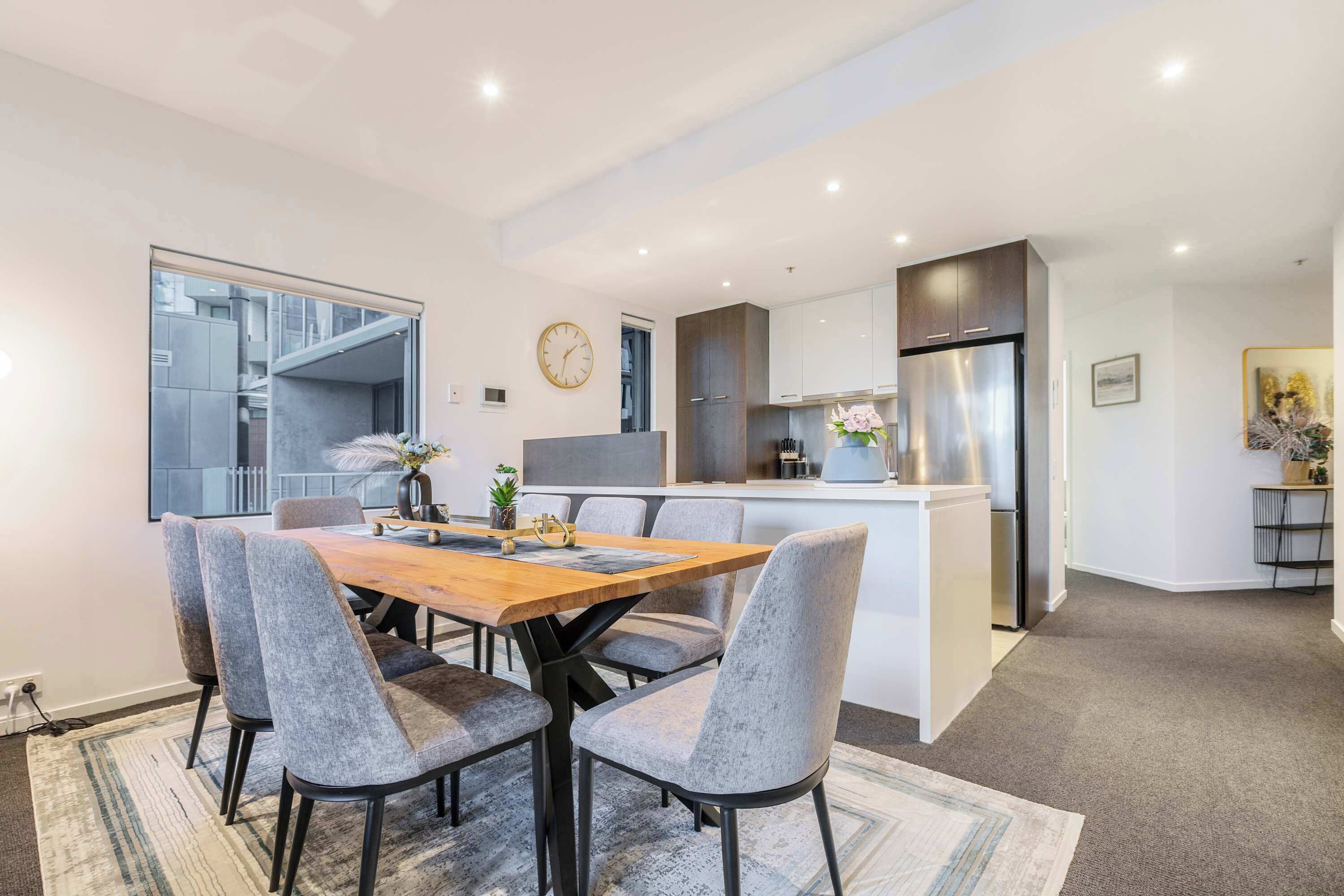 A modern kitchen with a central island, a dining area with grey upholstered chairs, and a large window.