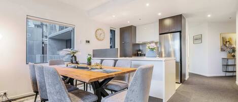 A modern kitchen with a central island, a dining area with grey upholstered chairs, and a large window.