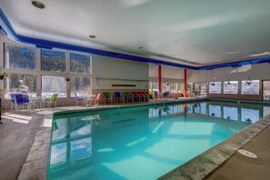 An indoor swimming pool with clear blue water, surrounded by colorful lounge chairs and large windows offering views of the outside.