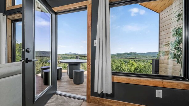 A modern room with a sliding glass door leading to an outdoor deck with a table and chairs, a view of a forested landscape, and a wooden ceiling.