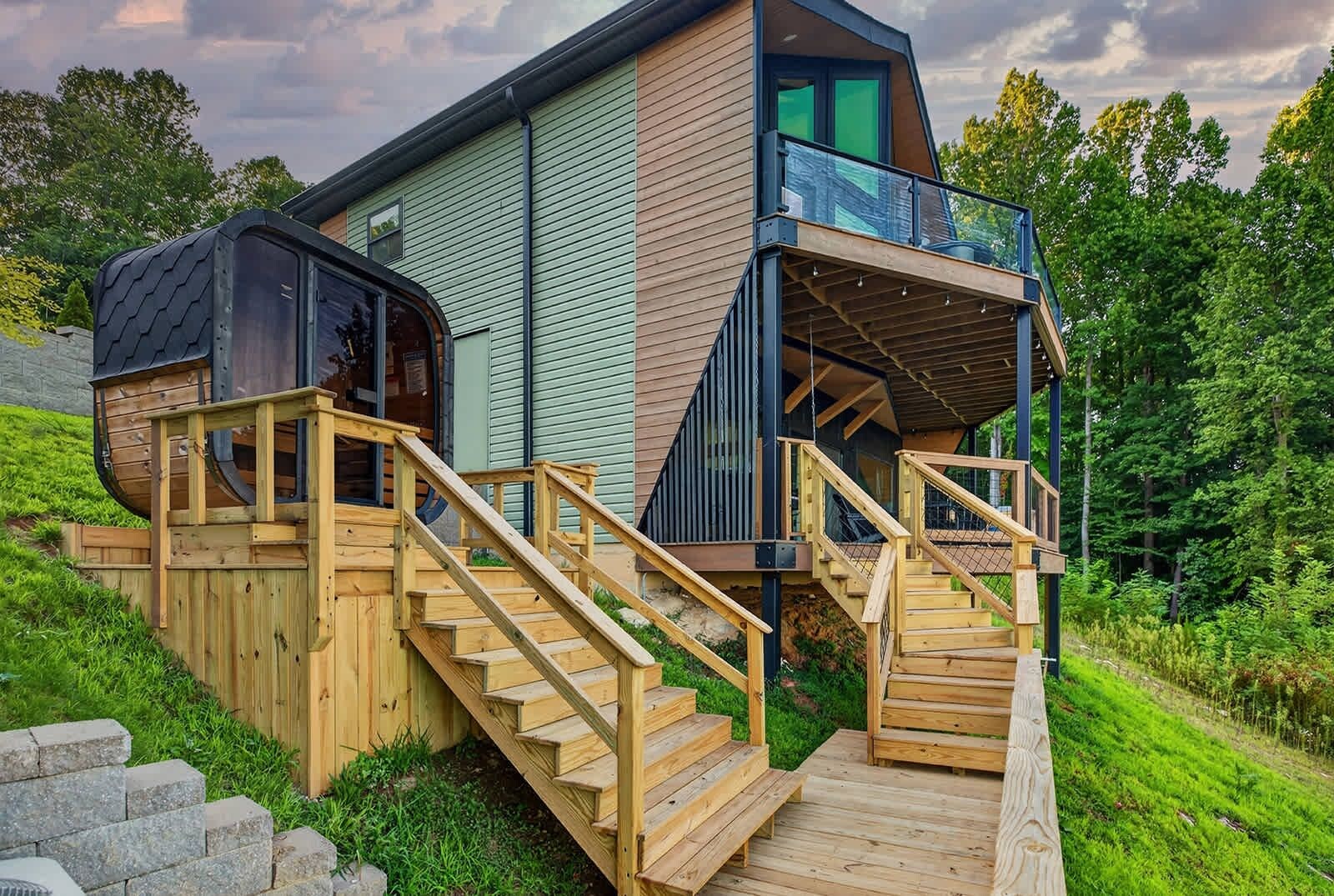 Une maison moderne dotée d’une terrasse en bois, d’un balcon en verre et d’un escalier menant à l’entrée.