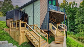 Une maison moderne dotée d’une terrasse en bois, d’un balcon en verre et d’un escalier menant à l’entrée.
