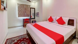 A hotel room with a bed, a desk, a chair, a ceiling fan, and a window with curtains. - HOTEL MAGADH REGENCY (Patna)