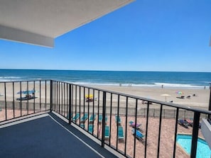 A balcony with a view of the beach, poolside chairs, and a swimming pool.