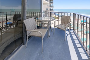 A balcony with wicker chairs and a table, overlooking a beach with umbrellas and people.