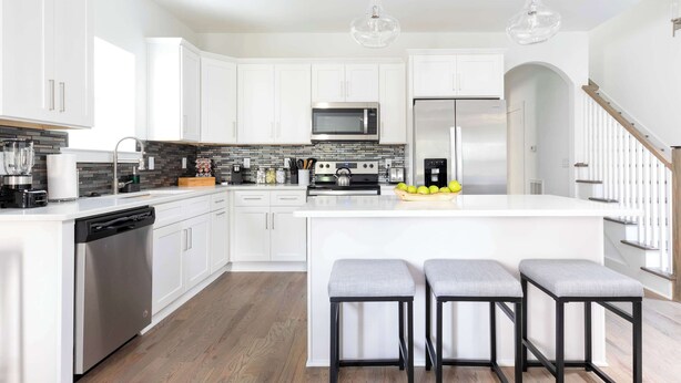 A modern kitchen with white cabinetry, a central island with bar stools, a stainless steel dishwasher, a microwave, and a refrigerator.
