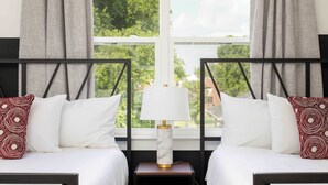 A hotel room with two beds, a nightstand with a lamp, and a window with a view of greenery and a bridge.