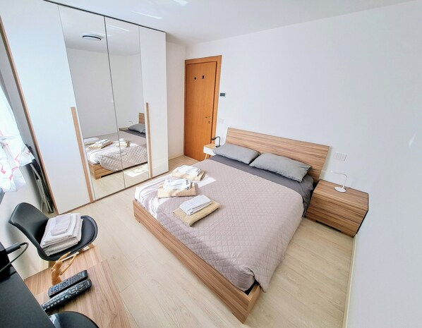 A modern hotel room with a large bed, a wooden headboard, a bedside table, a chair, and a mirror.