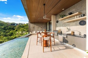 A modern outdoor kitchen with a dining area overlooking a pool and lush greenery. - 6 BR Architectural Masterpiece in Peninsula Papagayo (Nacascolo)