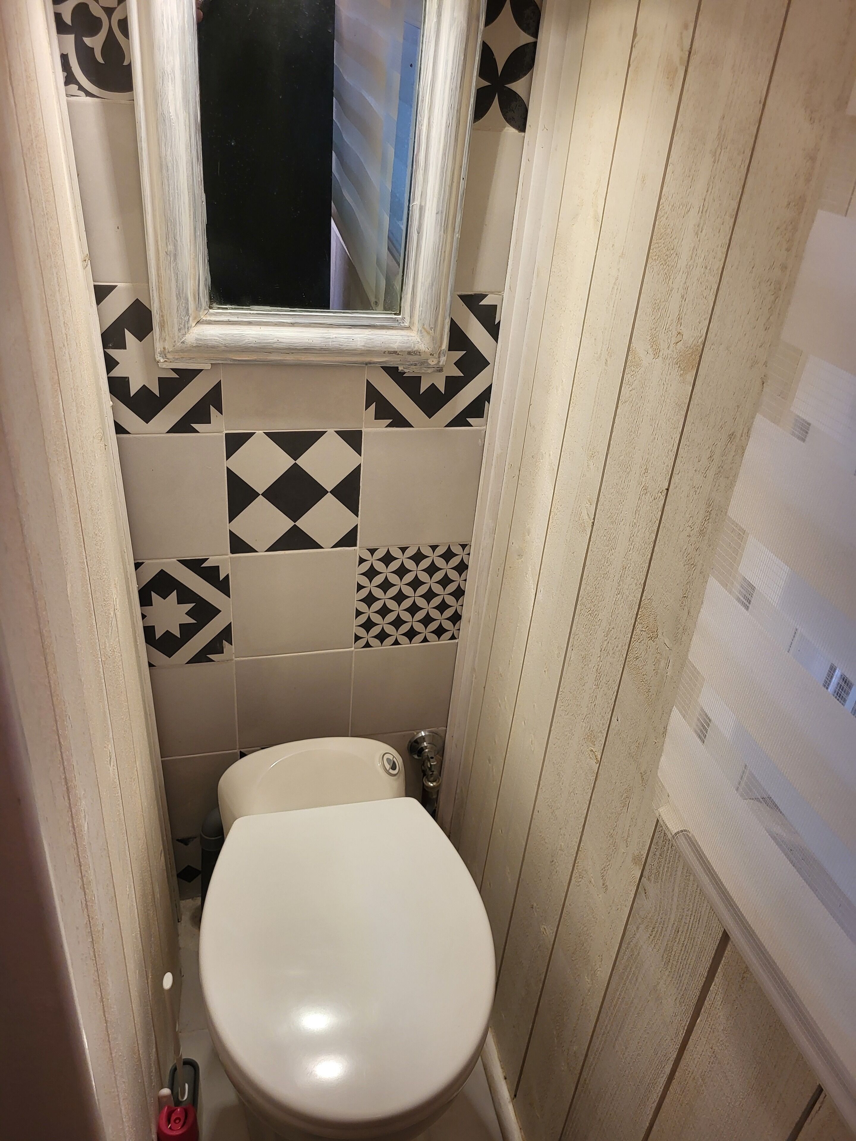 Ein kleines, schmales Badezimmer mit einer weißen Toilette, einem Fenster über dem Waschbecken und Wandfliesen mit Muster.