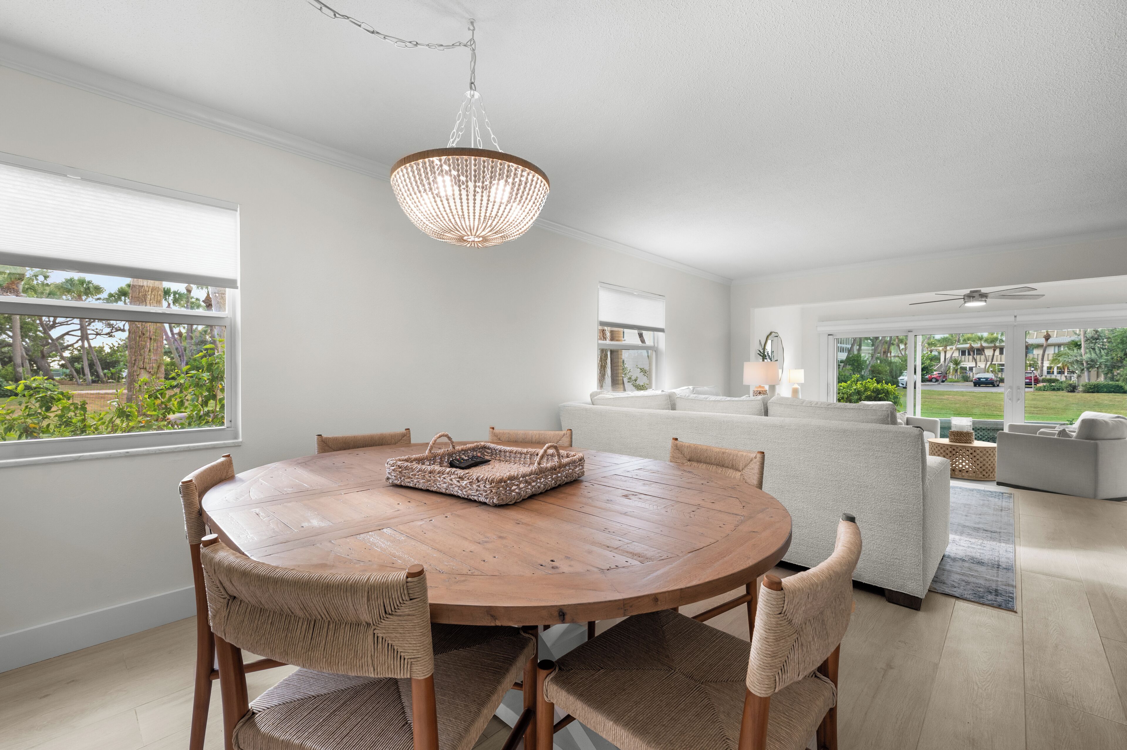 Een ruime woonkamer met een ronde houten eettafel, rieten stoelen, een witte bank en een groot raam met uitzicht op de omgeving.
