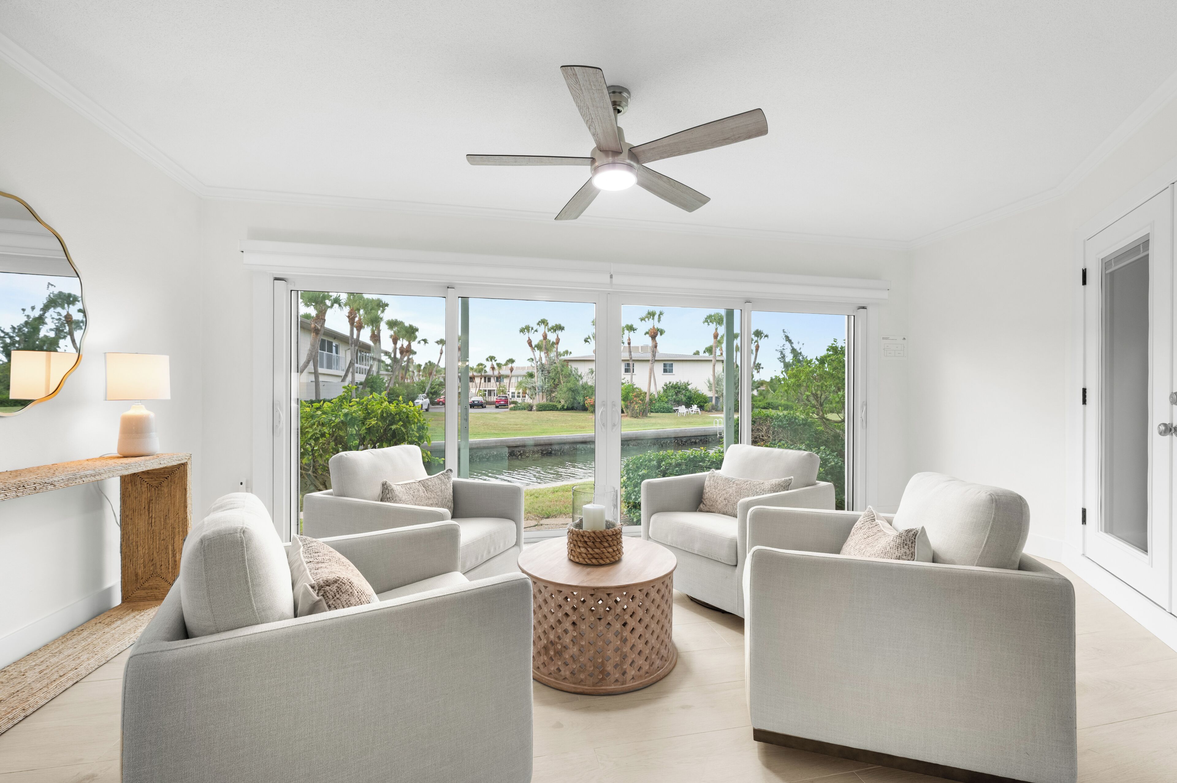 Un salon moderne doté d’un ventilateur de plafond, de grandes fenêtres et offrant une vue sur un lac et des palmiers.