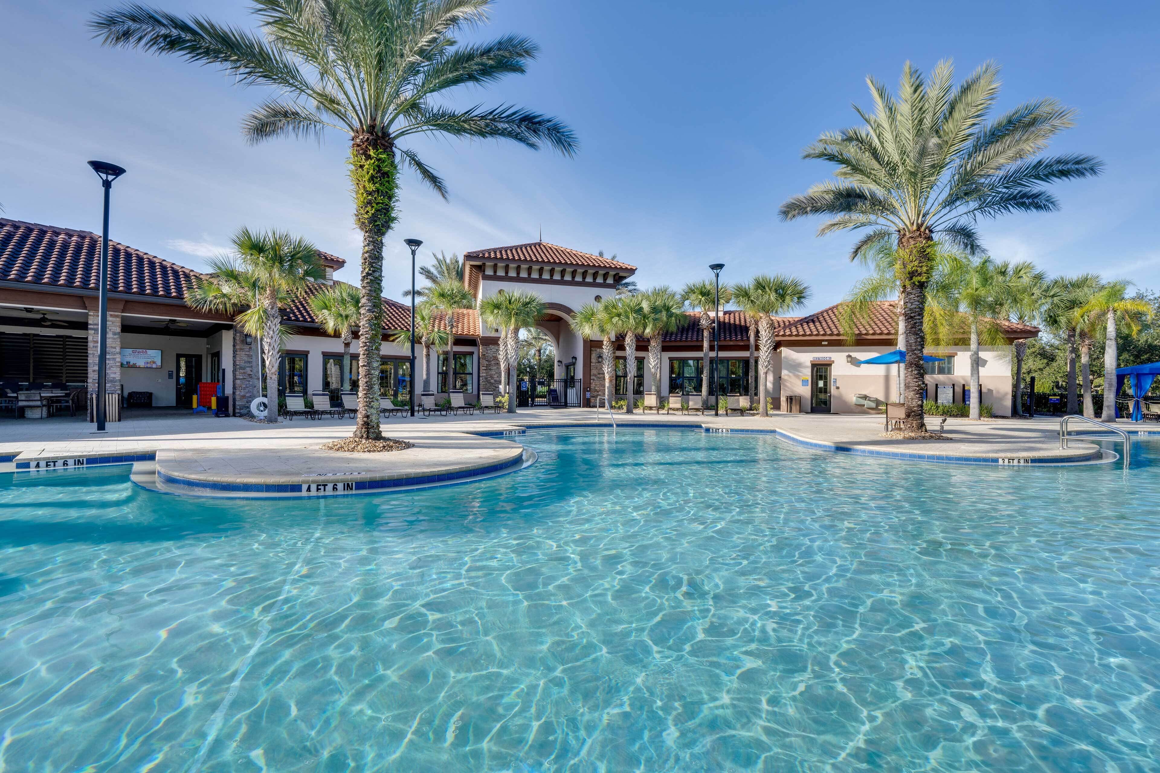 A clear swimming pool with palm trees and a building with a tiled roof in the background.