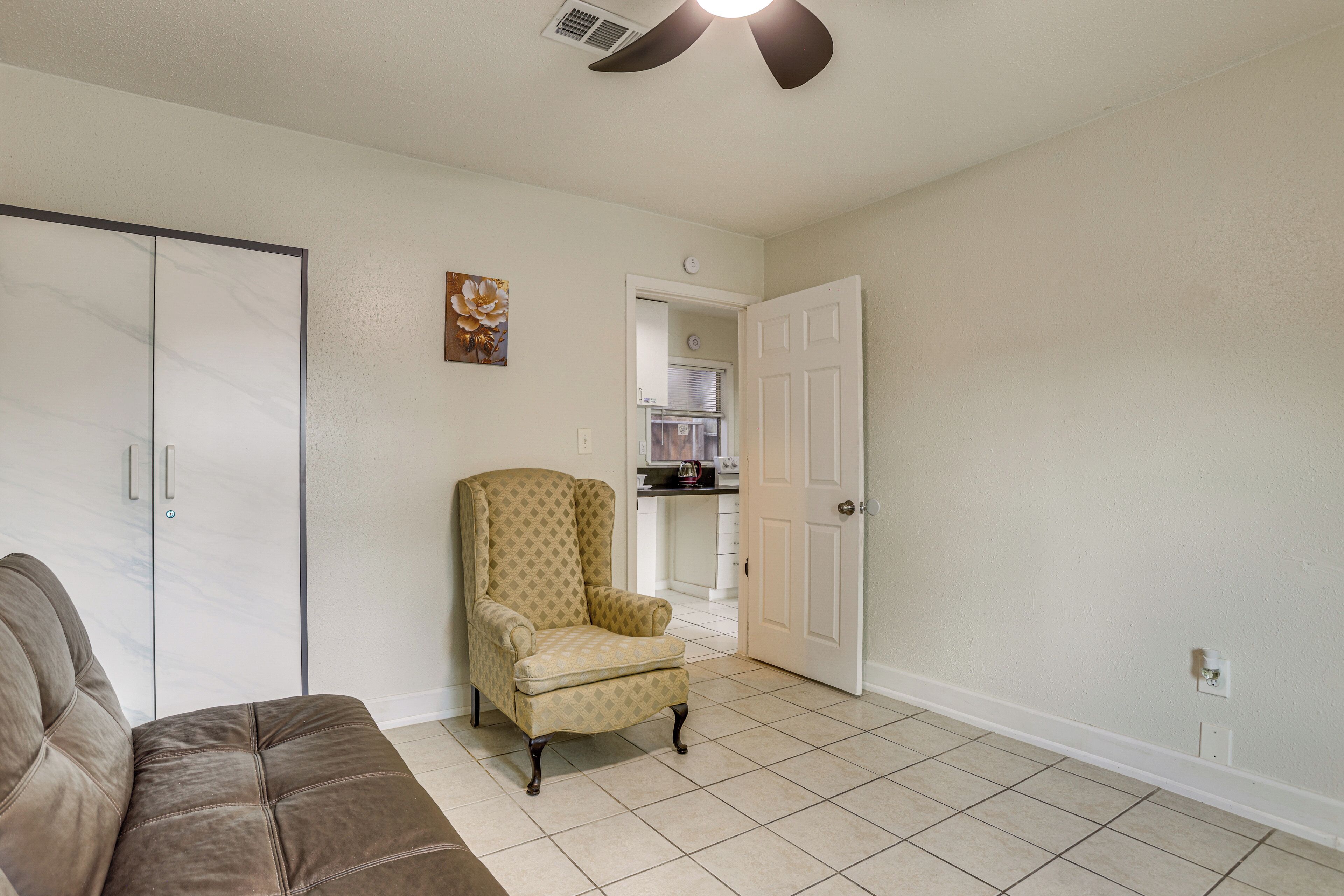 A living room with a brown sofa, a patterned armchair, a white door, and a kitchen in the background.