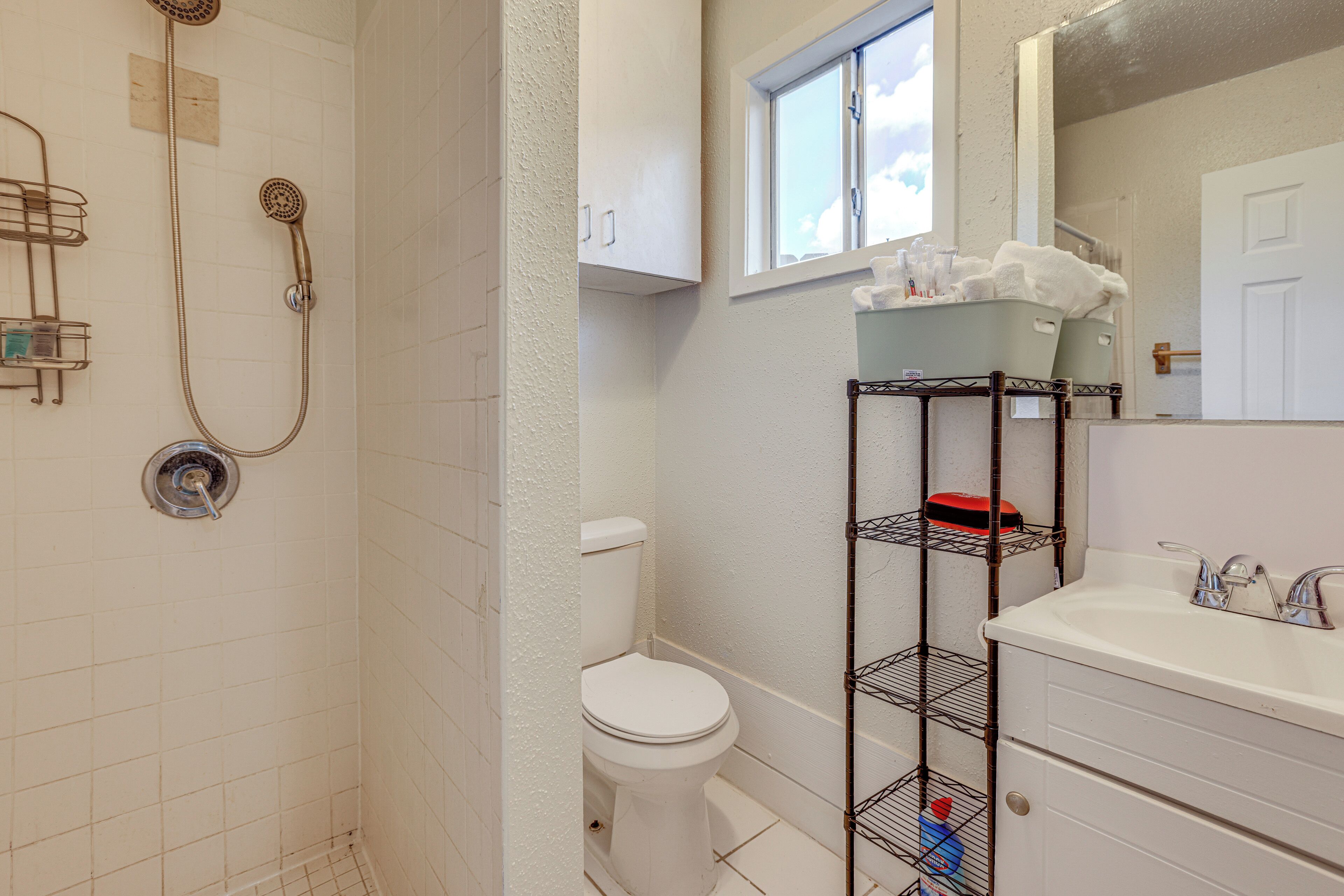 Une salle de bain avec une douche, des toilettes et un lavabo avec un miroir. On y trouve des étagères avec des articles de toilette et une fenêtre au-dessus des toilettes.