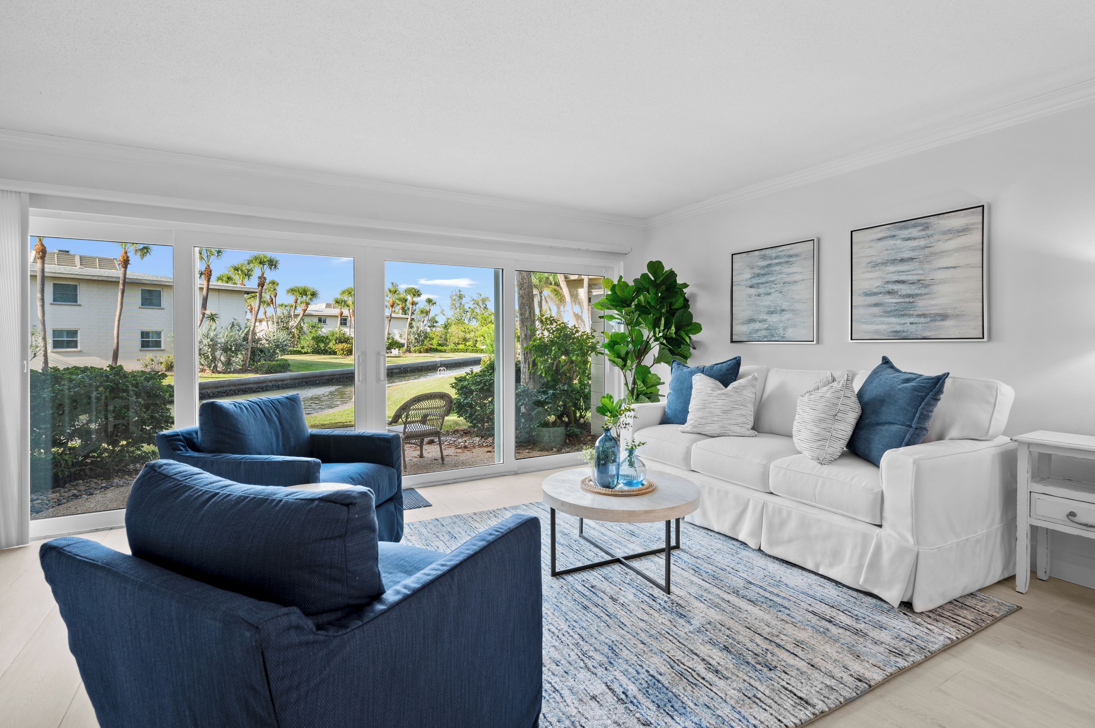 A modern living room with a white sofa, blue armchairs, and a coffee table. Large windows offer a view of a residential area with palm trees.