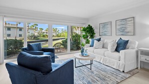 A modern living room with a white sofa, blue armchairs, and a coffee table. Large windows offer a view of a residential area with palm trees. - LBK Whitney Beach Retreat 1 (Longboat Key)