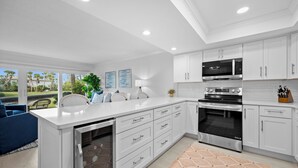 A modern kitchen with white cabinetry, a built-in microwave, and an oven. There is a dining area with a blue chair and a plant on the counter. - LBK Whitney Beach Retreat 1 (Longboat Key)