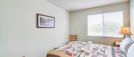 Uma cama bem arrumada com um edredom, um quadro na parede, uma janela com persianas e um abajur em uma mesa de cabeceira.