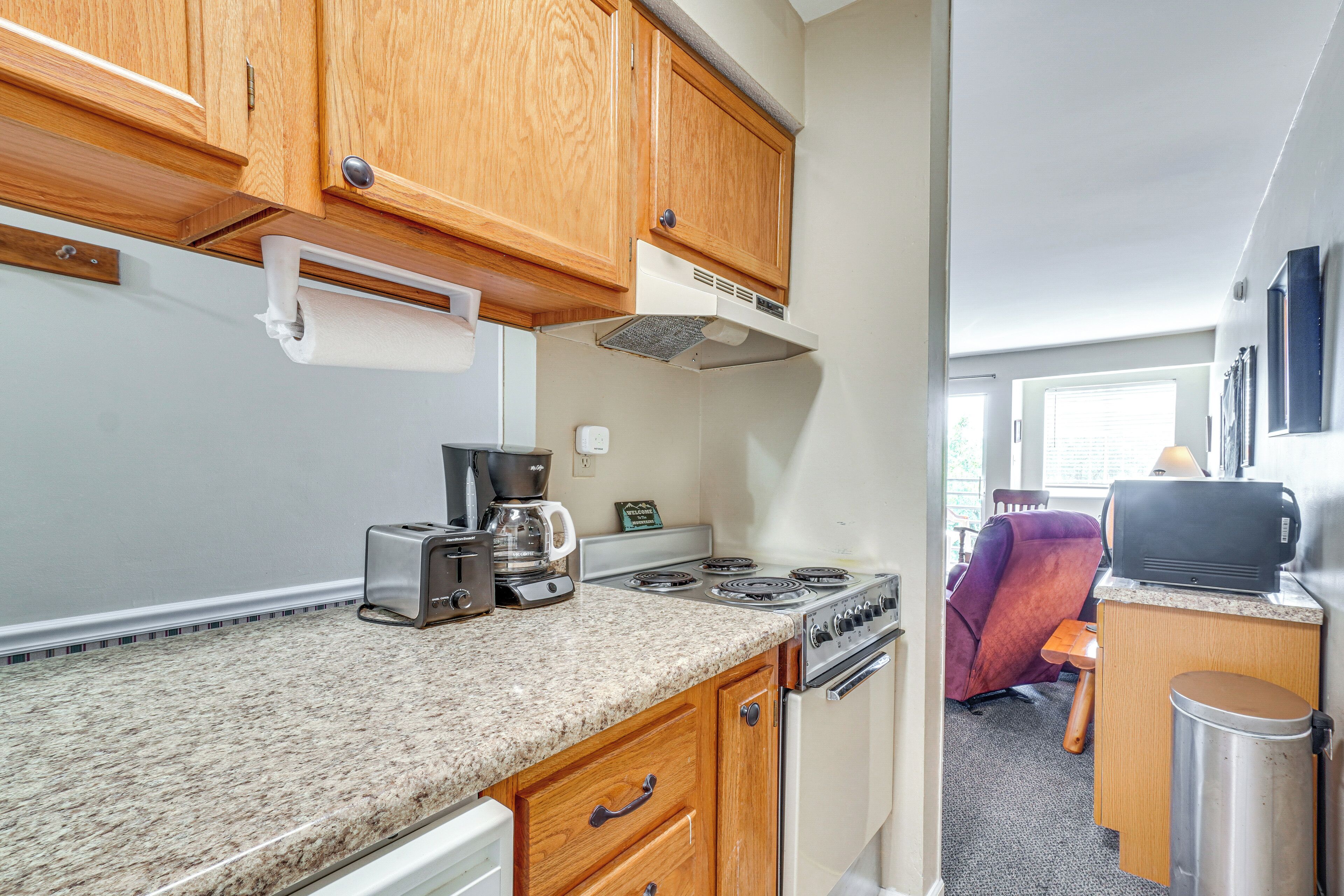 A compact kitchen with wooden cabinets, a gas stove, a coffee maker, and a trash can.