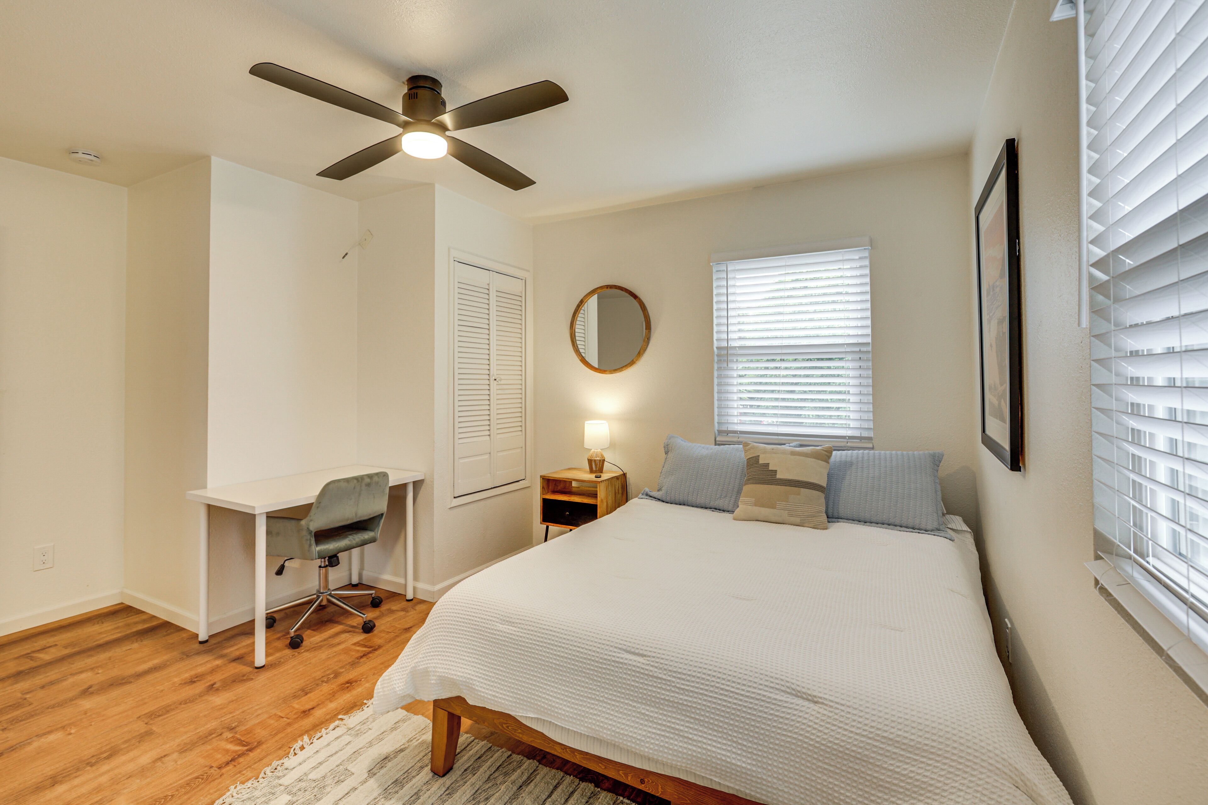 Une chambre à coucher comprenant un lit, un bureau, une chaise, un ventilateur de plafond, une lampe de chevet et une fenêtre avec des stores.