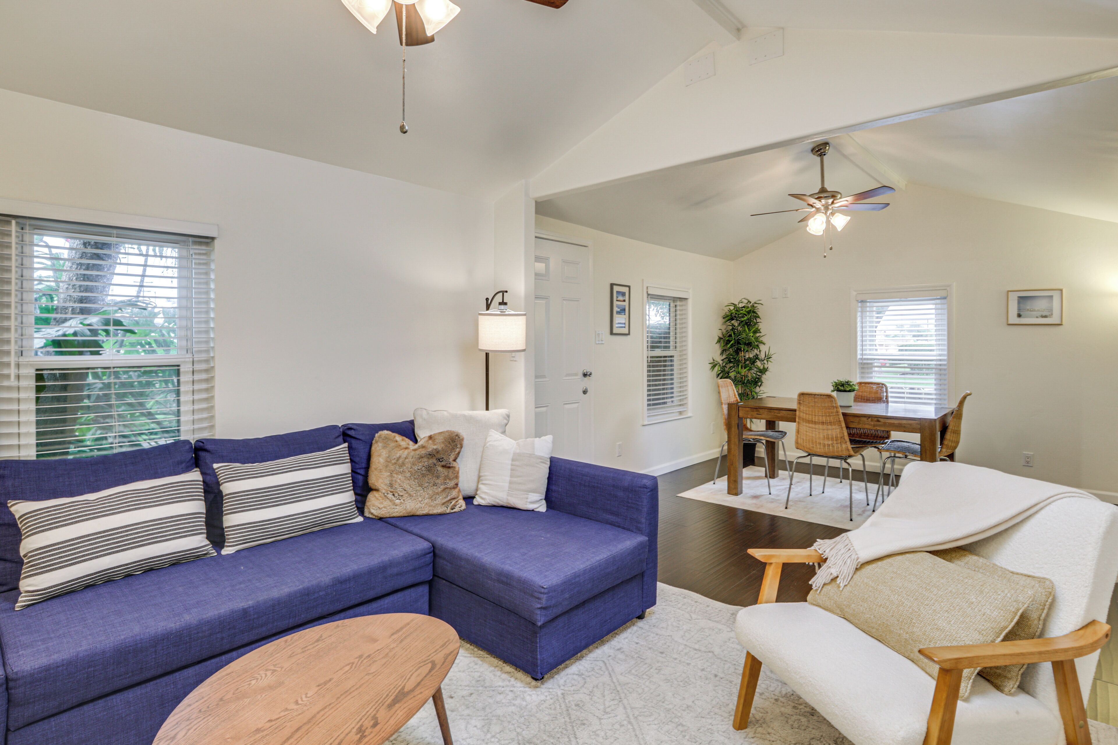 A living room with a blue sectional sofa, a wooden coffee table, and a white armchair. There is a dining area with a wooden table and chairs, and a ceiling fan.
