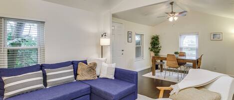 A living room with a blue sectional sofa, a wooden coffee table, and a white armchair. There is a dining area with a wooden table and chairs, and a ceiling fan.