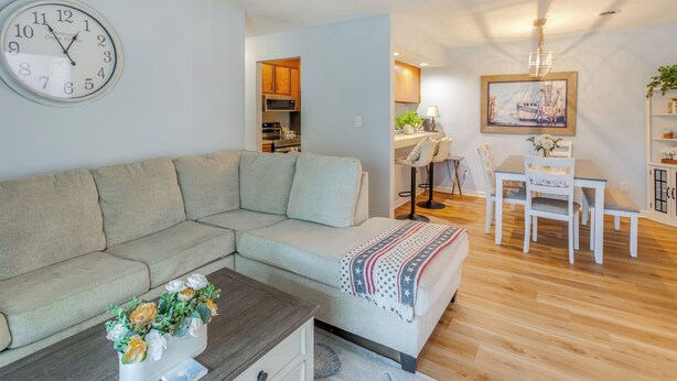 A living room with a sectional sofa, a coffee table with flowers, a dining area with a table and chairs, and a kitchen in the background. - 1/2 Mi to Shore: Condo w/ Balcony in Myrtle Beach! (Myrtle Beach)