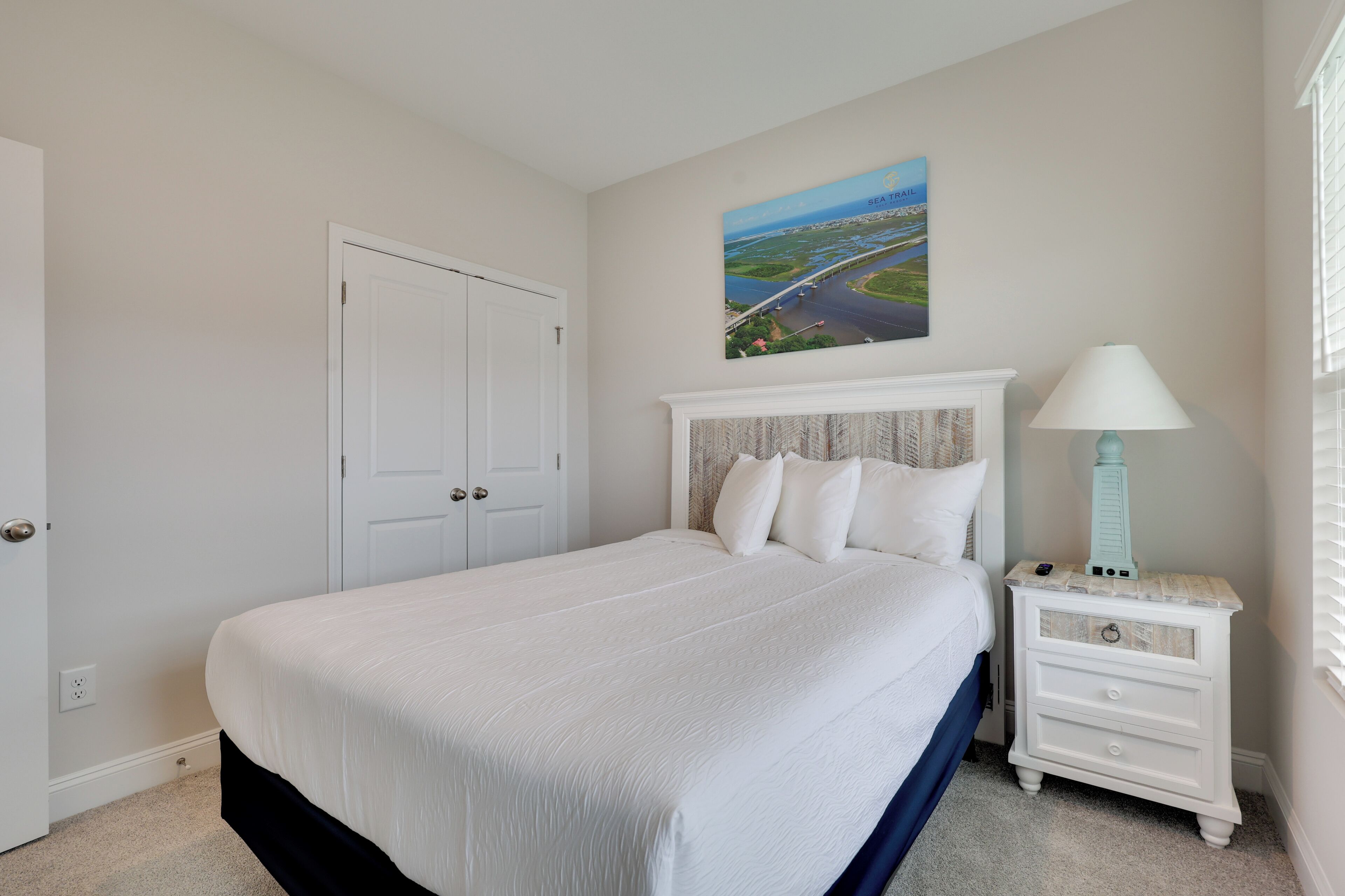 A bedroom with a large bed, a nightstand with a lamp, a door, and a painting on the wall.