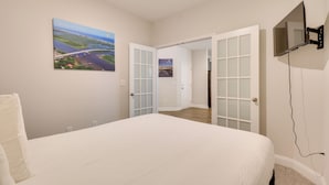 A bedroom with a large bed, a TV mounted on the wall, and a scenic view of a bridge and landscape through an open door. - 3 Mi to Sand & Surf: Ocean Isle Beach Retreat! (Ocean Isle Beach)