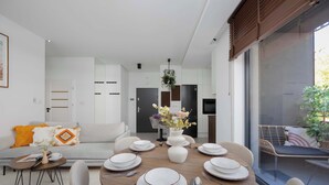 A modern dining area with a round table set for six, surrounded by chairs. A white vase with flowers is placed on the table, and there is a view of the kitchen and living area in the background.
