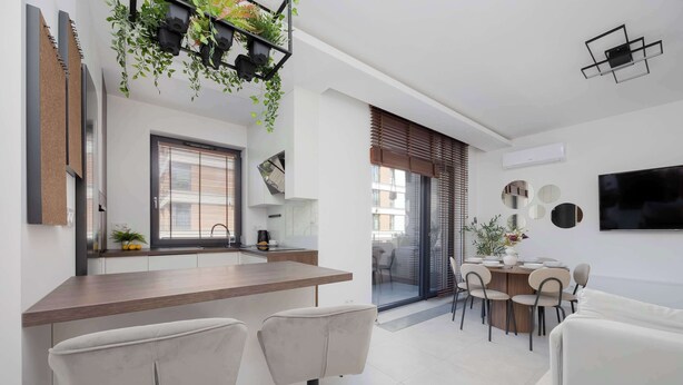 A modern kitchen with a wooden countertop and bar stools, a dining area with a table and chairs, and a living space with a sofa and a TV mounted on the wall.