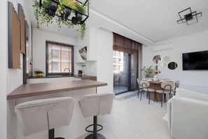 Una cocina moderna con encimera de madera y taburetes de bar, un comedor con mesa y sillas, y una sala de estar con sofá y televisor montado en la pared.