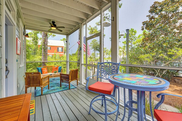 A covered outdoor deck with wooden furniture, a round table with a colorful glass top, and a view of trees and buildings in the background. - Poolside Fun + Ocean Strolls: Port St Joe Retreat! (Port St. Joe)