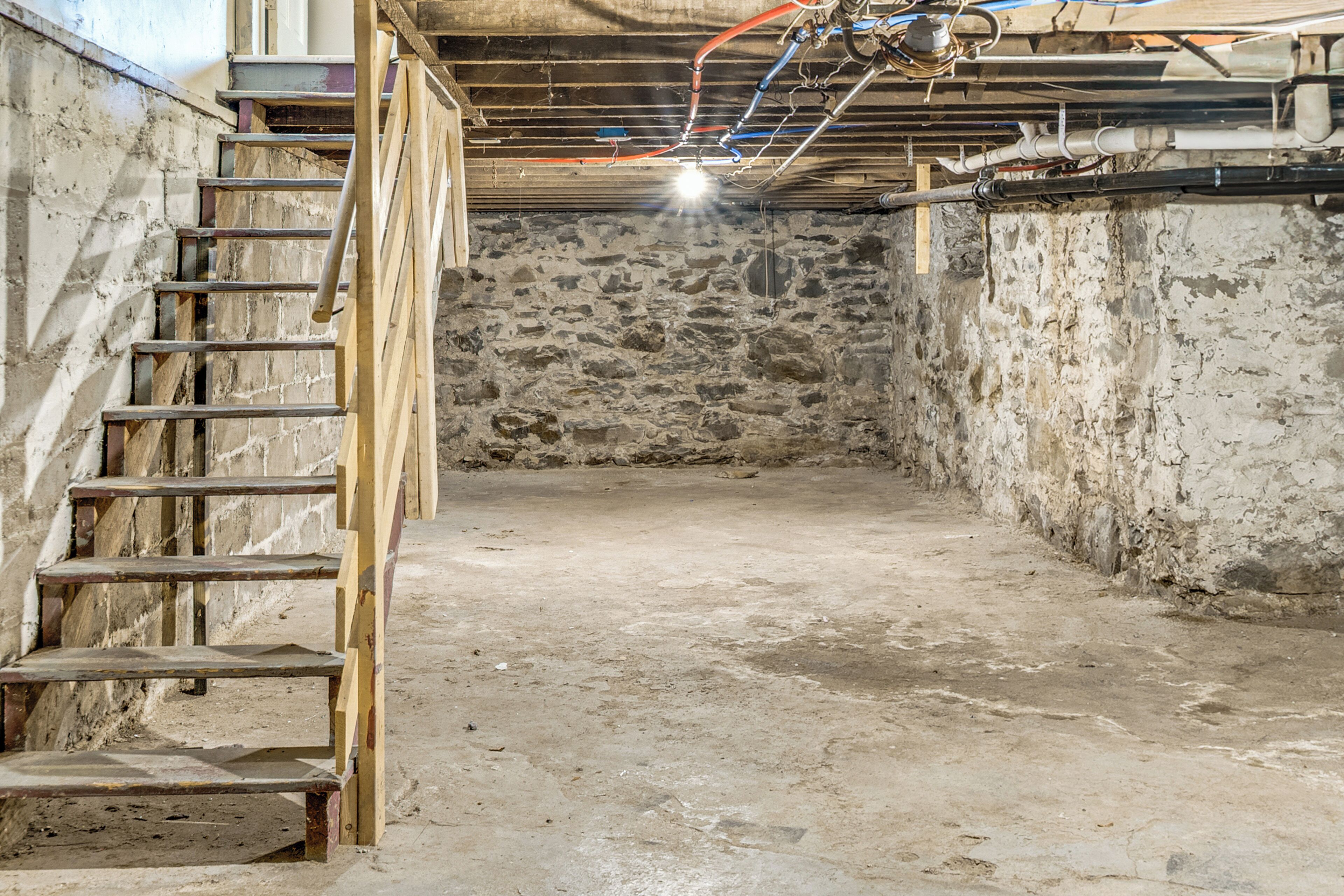 A basement with a wooden staircase leading up, a stone wall, and visible plumbing.