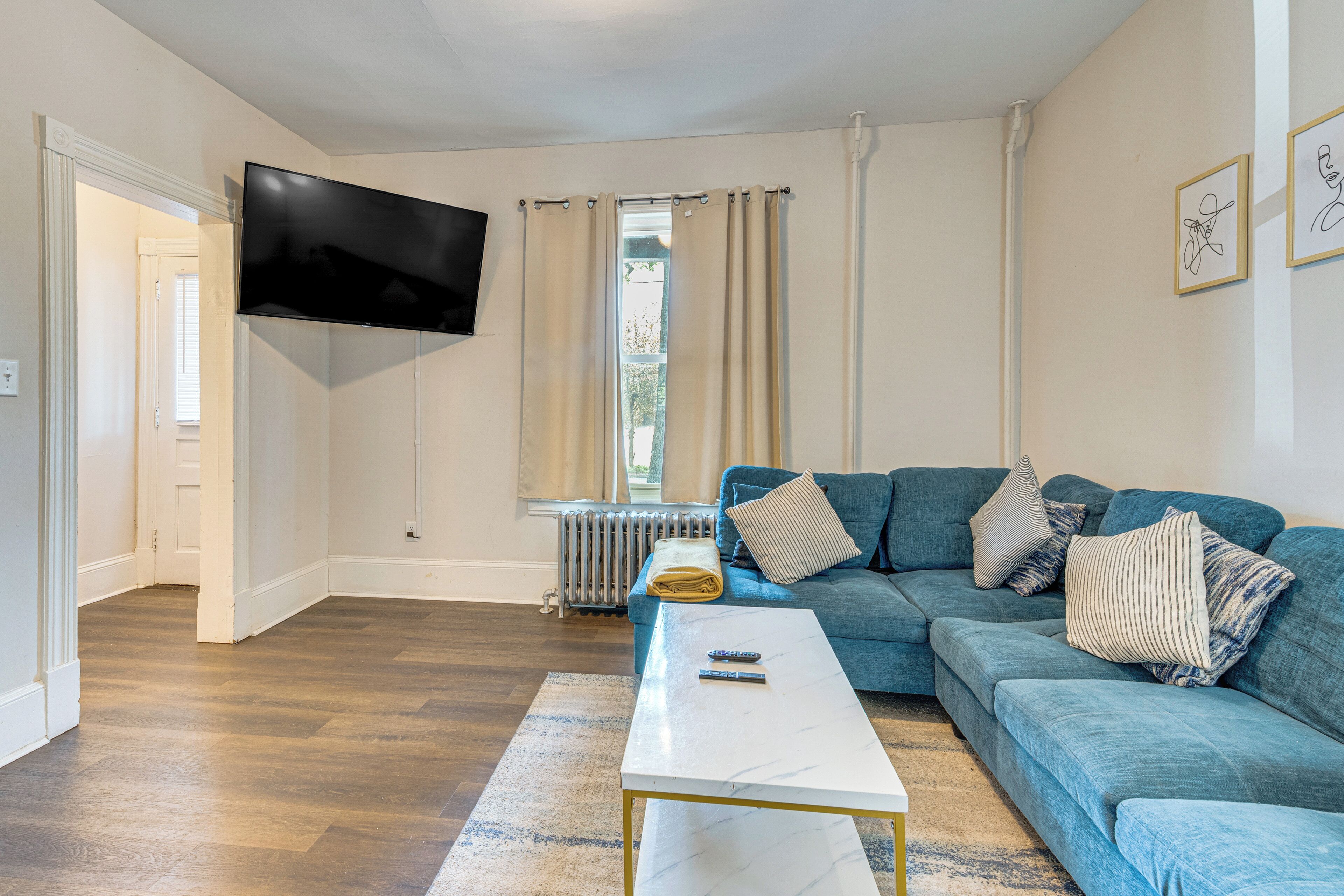 A living room with a blue sofa, a white coffee table, a flat-screen TV mounted on the wall, and a window with beige curtains.