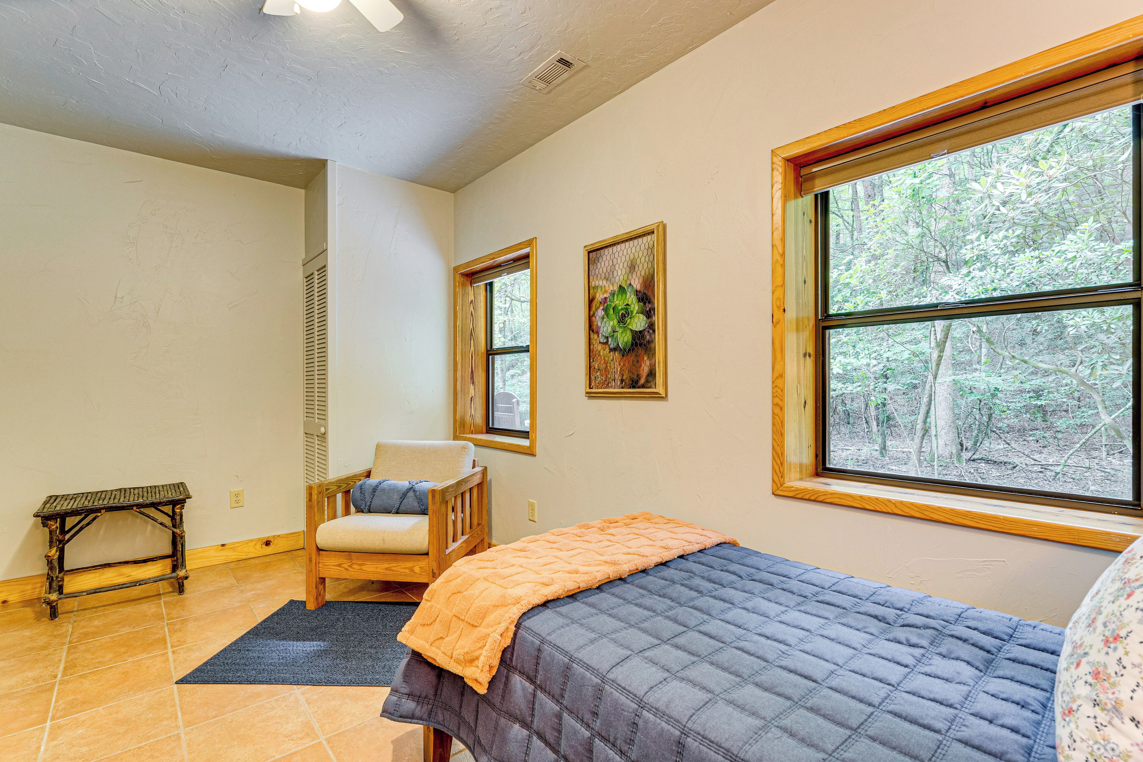 A bedroom with a bed, a chair, a small table, and two windows with views of trees.