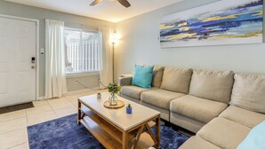 A living room with a beige sofa, a wooden coffee table, a ceiling fan, a painting on the wall, and a window with curtains. - Steps to Park: Dog-friendly Condo in Conyers! (Conyers)
