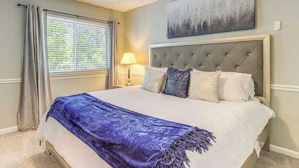 A bedroom with a large bed, a headboard, a window with blinds, a lamp, and a painting on the wall. - Steps to Park: Dog-friendly Condo in Conyers! (Conyers)