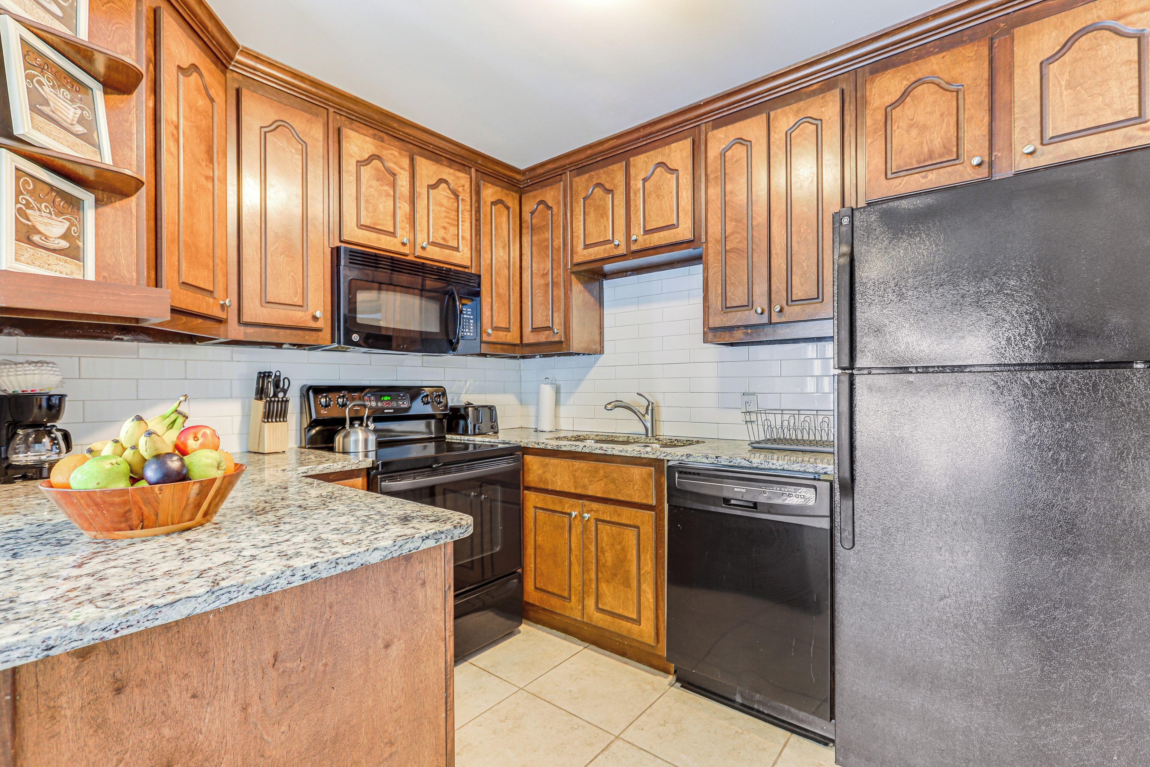 A kitchen with wooden cabinets, a black refrigerator, a microwave, a dishwasher, and a granite countertop with a fruit bowl.