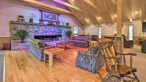 Ein gemütliches Wohnzimmer im Loft-Stil mit Kamin aus Stein, Holzbalken und vielfältigen Sitzgelegenheiten, darunter ein Karosobereich, ein gemustertes Sessel und ein Schaukelstuhl.