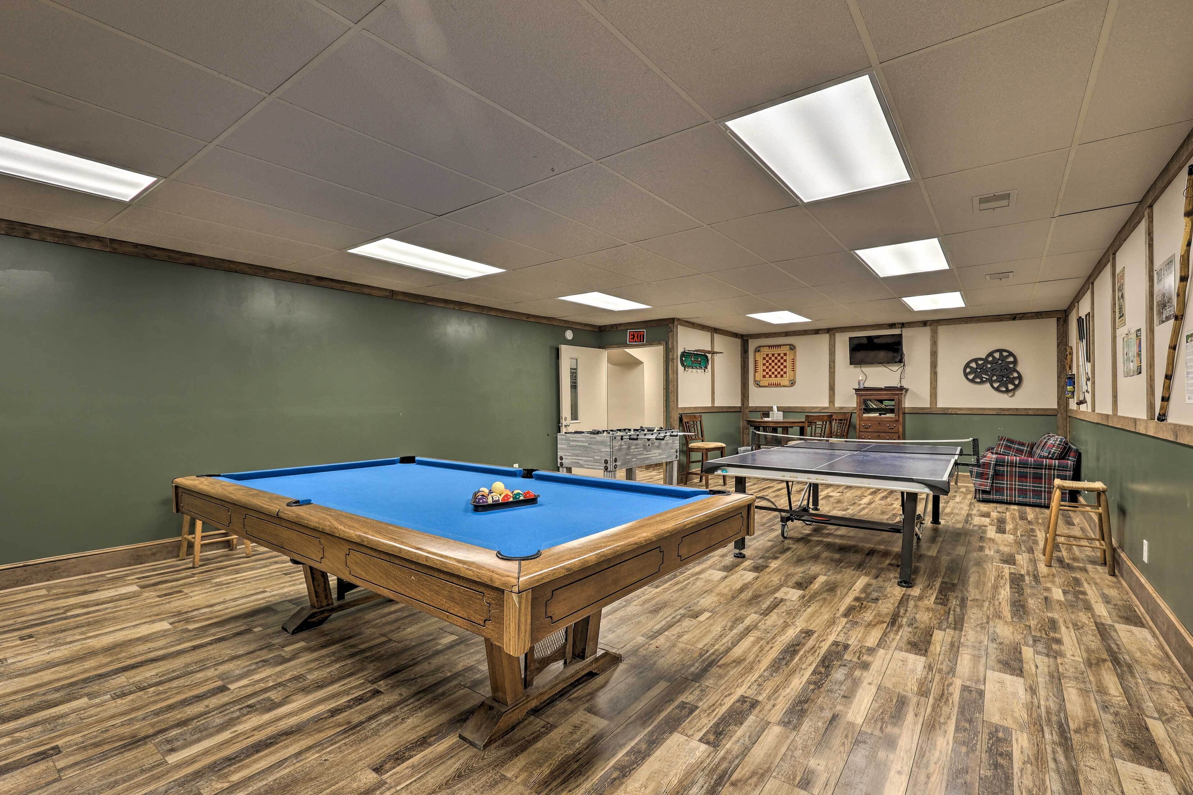 Una mesa de billar con superficie de fieltro azul y estructura de madera, una mesa de ping pong y una mesa de fútbol de mesa en una habitación con suelo de madera y paredes verdes.