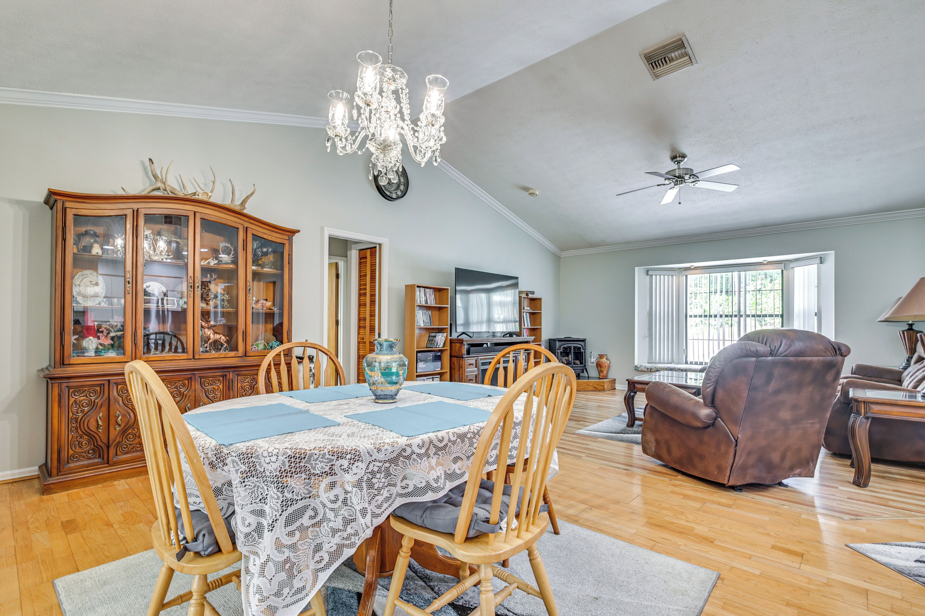 A spacious living room with a dining area, wooden furniture, a chandelier, and a ceiling fan.