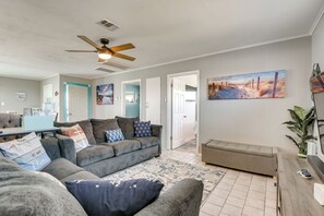 Un salon spacieux avec un ensemble de canapés gris, un ventilateur de plafond et un tableau sur le thème de la plage accroché au mur.