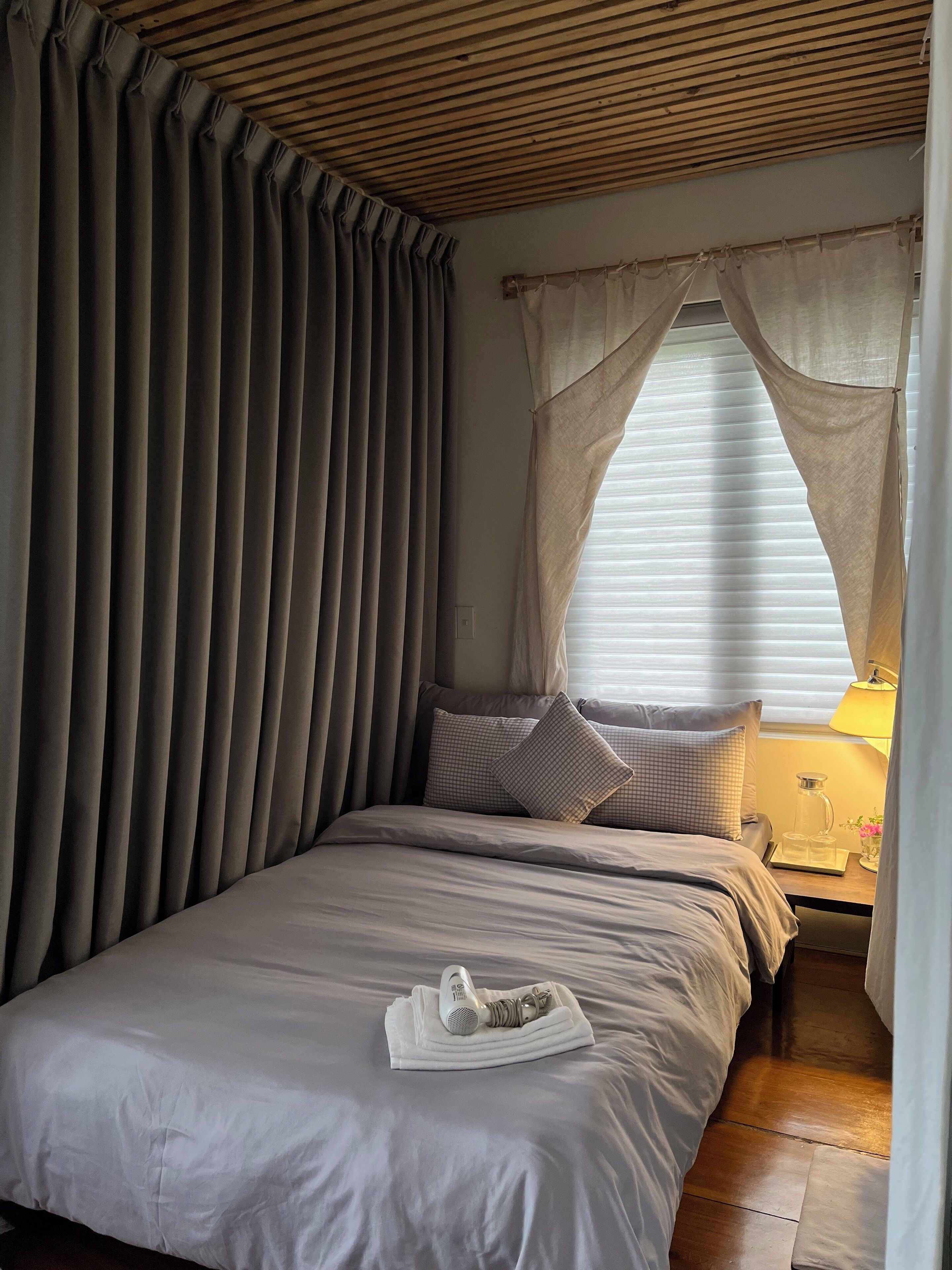 A neatly made bed with a pillow, a bedside table with a lamp, and a window with curtains.