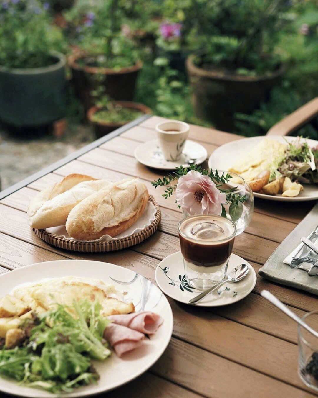 A wooden table set with plates of food, including bread, salad, and a cup of coffee, in an outdoor setting with potted plants in the background.