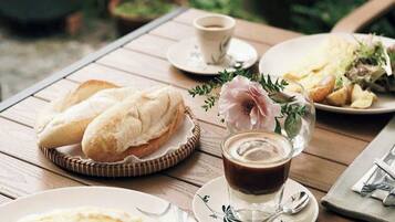 A wooden table set with plates of food, including bread, salad, and a cup of coffee, in an outdoor setting with potted plants in the background.