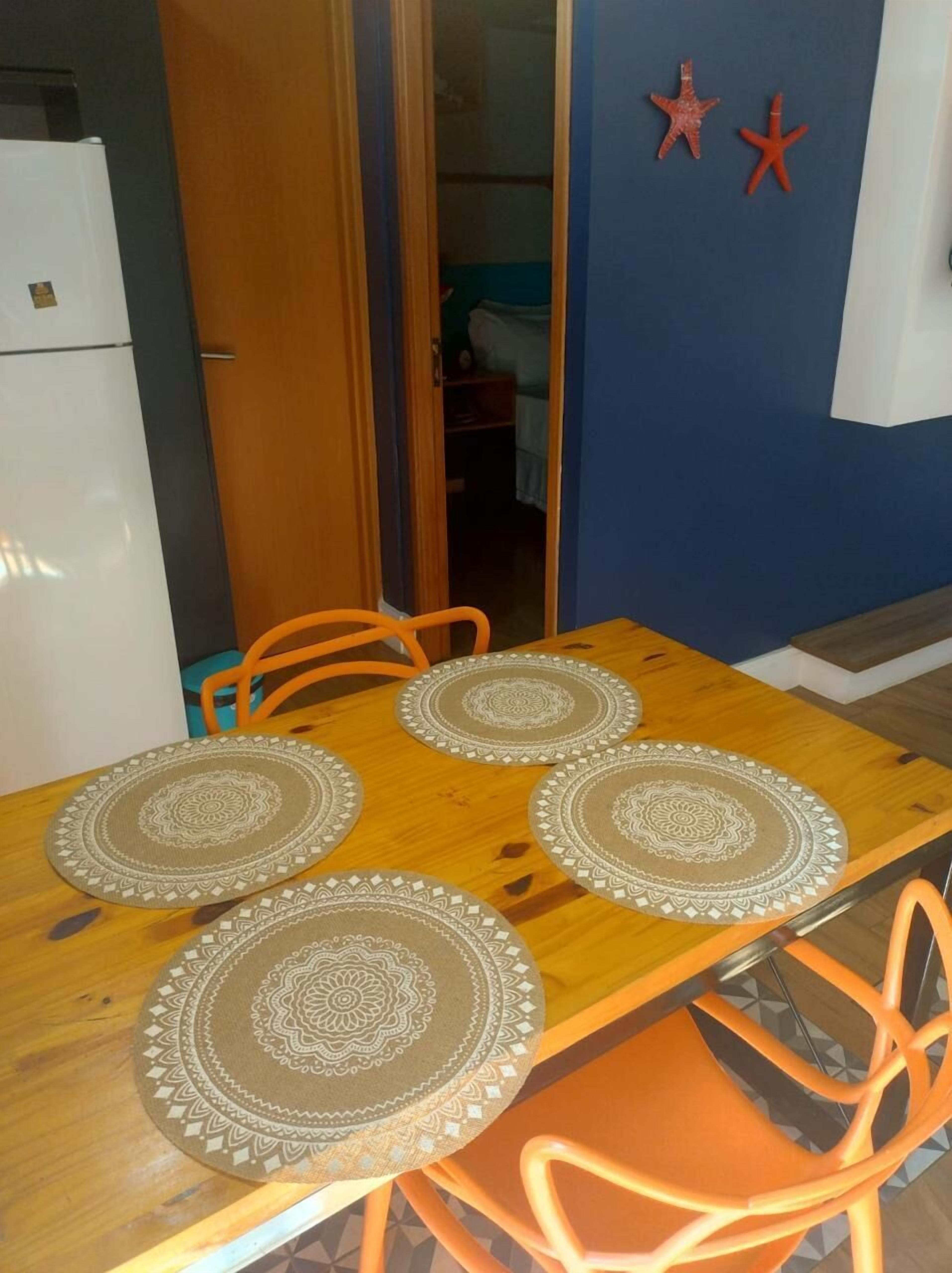 Una mesa de comedor con cuatro individuales, sillas color naranja y un refrigerador al fondo.