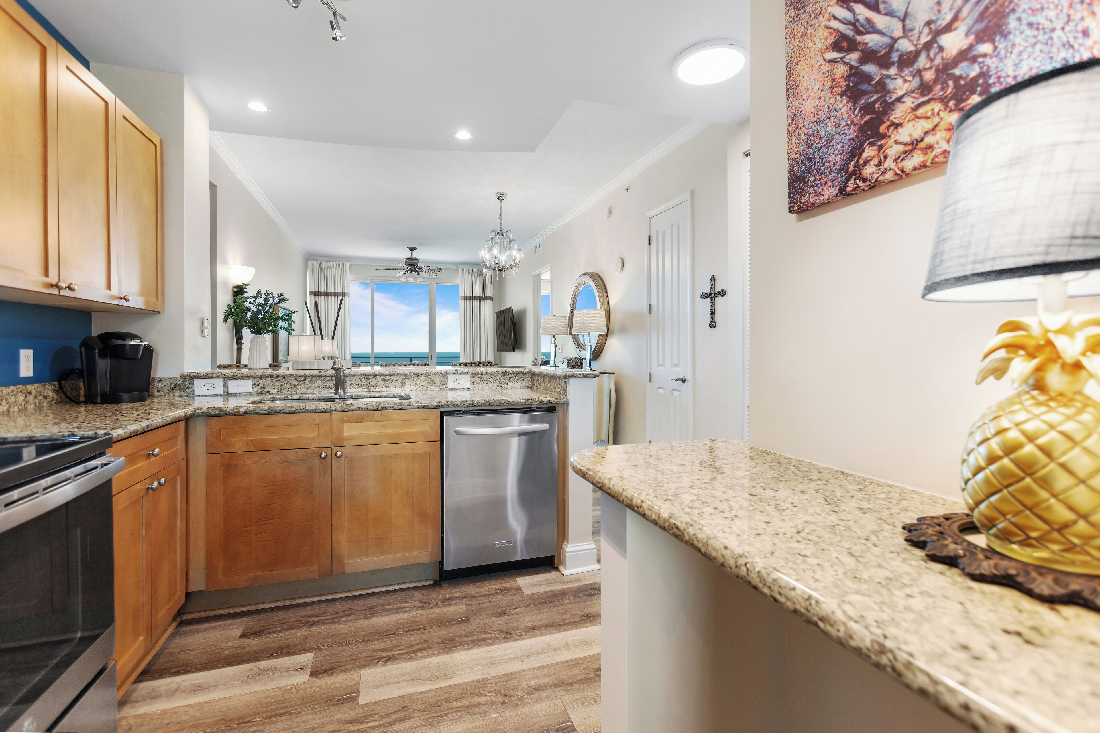 A kitchen with wooden cabinets, a stainless steel dishwasher, granite countertops, and a view of the ocean.