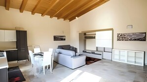 A modern kitchen with white cabinets and a black countertop. There is a white sofa and a dining table with chairs. The room has a high ceiling with wooden beams. - House Lumia White Zagara (Sicilia)